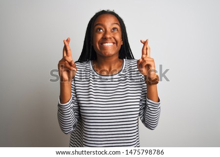 Young african american woman wearing striped t-shirt standing over isolated white background gesturing finger crossed smiling with hope and eyes closed. Luck and superstitious concept.