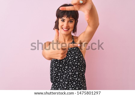 Young beautiful woman wearing black floral dress standing over isolated pink background smiling making frame with hands and fingers with happy face. Creativity and photography concept.