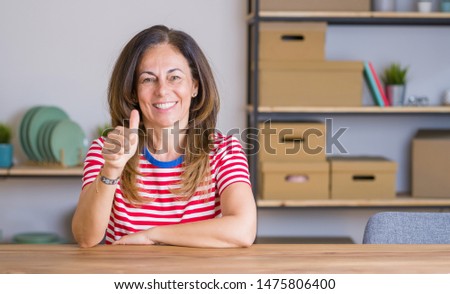 Middle age senior woman sitting at the table at home doing happy thumbs up gesture with hand. Approving expression looking at the camera showing success.