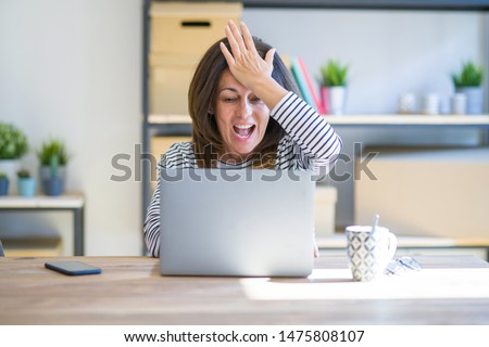 Middle age senior woman sitting at the table at home working using computer laptop surprised with hand on head for mistake, remember error. Forgot, bad memory concept. Royalty-Free Stock Photo #1475808107