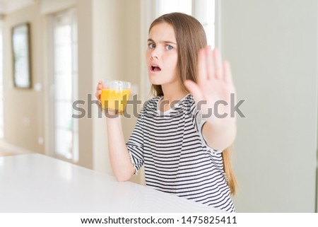 Beautiful young girl kid drinking a glass of fresh orange juice with open hand doing stop sign with serious and confident expression, defense gesture