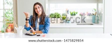 Wide angle picture of beautiful young woman eating delivery sushi smiling with happy face looking and pointing to the side with thumb up.