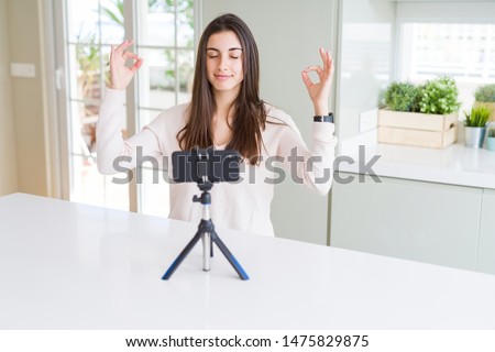 Beautiful young woman recording selfie video with smartphone webcam relax and smiling with eyes closed doing meditation gesture with fingers. Yoga concept.