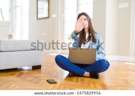 Beautiful young woman sitting on the floor with crossed legs using laptop with open hand doing stop sign with serious and confident expression, defense gesture