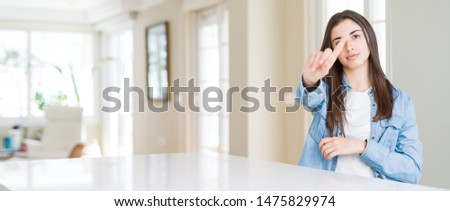 Wide angle picture of beautiful young woman sitting on white table at home Pointing with finger up and angry expression