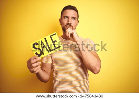 Young man holding sale advertising poster board over yellow isolated background serious face thinking about question, very confused idea
