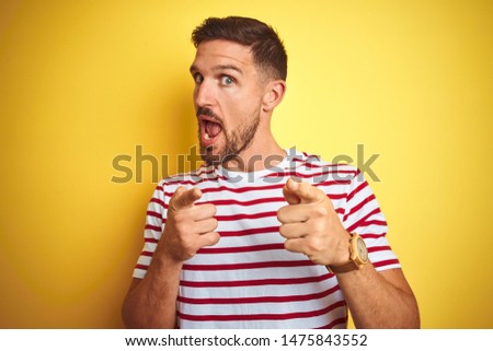 Young handsome man wearing casual red striped t-shirt over yellow isolated background pointing fingers to camera with happy and funny face. Good energy and vibes.
