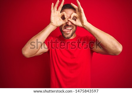 Young handsome man wearing casual t-shirt over red isolated background doing ok gesture like binoculars sticking tongue out, eyes looking through fingers. Crazy expression.