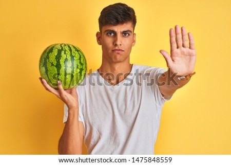 Young indian shopkeeper man holding watermelon standing over isolated yellow background with open hand doing stop sign with serious and confident expression, defense gesture