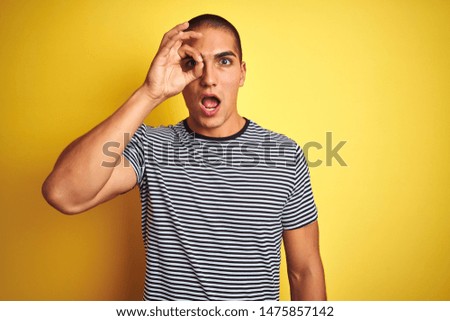 Young handsome man wearing striped t-shirt over yellow isolated background doing ok gesture shocked with surprised face, eye looking through fingers. Unbelieving expression.