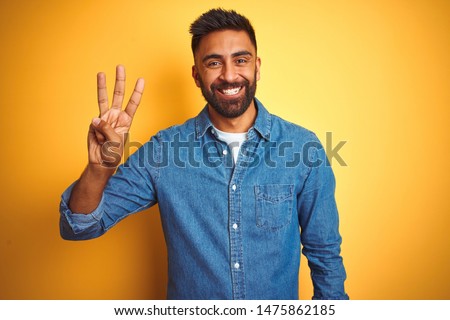 Young indian man wearing denim shirt standing over isolated yellow background showing and pointing up with fingers number three while smiling confident and happy. Royalty-Free Stock Photo #1475862185