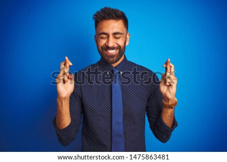Young indian businessman wearing elegant shirt and tie standing over isolated blue background gesturing finger crossed smiling with hope and eyes closed. Luck and superstitious concept.