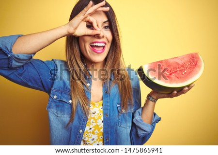 Young beautiful woman eating fresh healthy watermelon slice over yellow background with happy face smiling doing ok sign with hand on eye looking through fingers