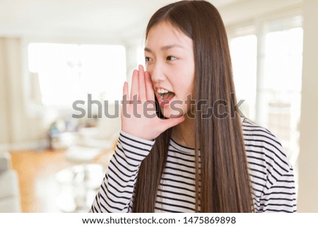 Beautiful Asian woman wearing stripes sweater over living room background shouting and screaming loud to side with hand on mouth. Communication concept.