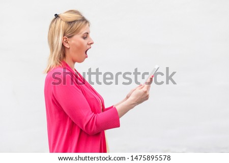No way! Side view portrait of unbelievable young woman in pink blouse standing and watching suprised video on her tablet with shocked face. Indoor, isolated, studio shot, white background