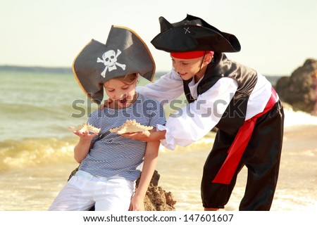 Cute little boy in a pirate costume and a little girl in a hat with a skeleton symbol of piracy are sitting on a large rock on the beach on a summer day