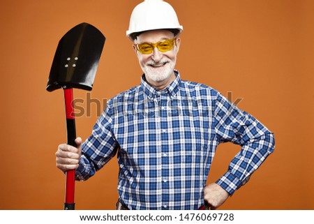 Studio image of joyful male construction employee on retirement wearing protective helmet and yellow goggles, using shovel for digging, posing isolated against blank copyspace wall background