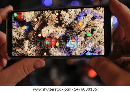 Elderly man holds in his hands new model of  smartphone and take pictures of garlands and Christmas lights on New Year tree. Christmas Eve