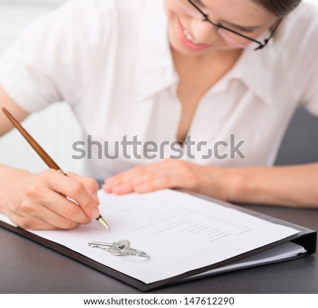 Female realtor signing mortgage contract in the office. Pretty smiling woman real estate seller reading partnership agreement with a house key lying on it.   