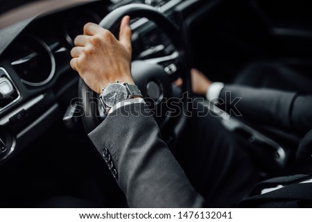 Close up top view of  man's watch in black suit keeping hand on the steering wheel while driving a luxury car. Royalty-Free Stock Photo #1476132041