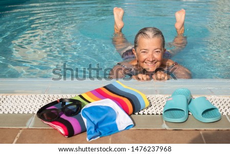 an old lady, amused, swimming to keep fit. outdoor pool with clear water and sunny day. Caucasian and smiling. Colorful towel on board