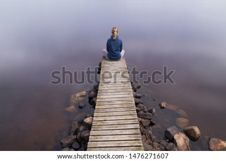 Young woman enjoying life sitting in yoga position on an old wooden pier looking into eternity over a calm water surface.