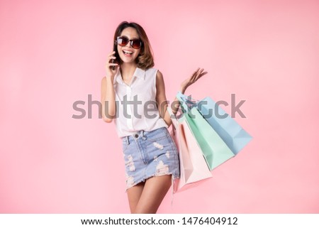 Asian pretty young woman walking over pink  pastel background. She smiling and  holding shopping bags. she nice-looking lovely attractive shine She holding mobile.
