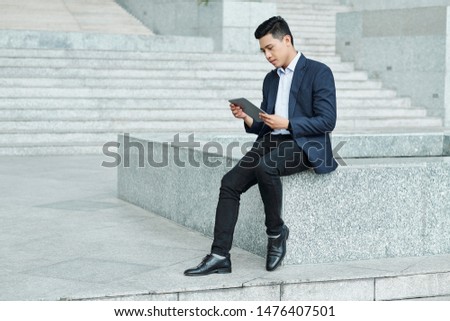 Young successful Vietnamese businessman sitting outdoors and reading news on screen of tablet computer