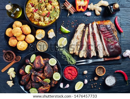 set of caribbean dishes, jerk pork belly, chicken curry, fried dumplings, roasted chicken thighs and drumsticks on plates on a black wooden table with spices and herbs, view from above, flat lay Royalty-Free Stock Photo #1476457733