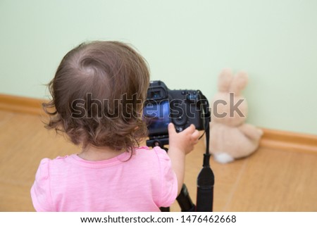 Cute funny beautiful baby in a pink T-shirt photographs her bunny. Close-up of the back of a baby girl and hand holding a DSLR. Concept novice beginner photographer, first steps.