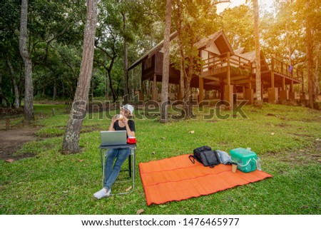 Pretty Asian women sitting on chair and eating bread and drinking coffee in vacation time, blurred homestay and forest on hill background, notebook and red coffee cup on table, bagpack and basket food