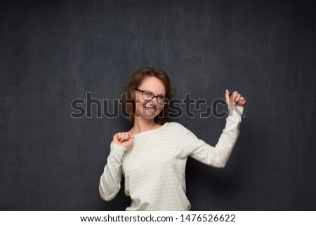 Studio waist-up portrait of happy caucasian fair-haired young woman with glasses, wearing jumper, raising hands while dancing, smiling broadly and looking cheerfully aside, over gray background