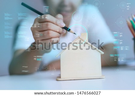 Happy Asian woman using pencil to draw with wooden house piggy bank and Cyber technology HUD GUI user interface metaphor design and decoration the house select focus shallow depth of field