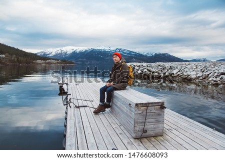 Traveler man with a yellow backpack wearing a red hat sitting on wooden pier on the background of mountain and lake. Space for your text message or promotional content. Travel lifestyle concept. A