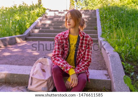 Tween tourist girl wearing casual clothes with backpack sitting on stairs in public garden. Beautiful look, summer or autumn street outfit. Urban vacation, teenage fashion concept