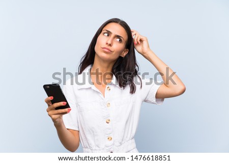 Young woman with a mobile phone over isolated blue wall having doubts and with confuse face expression Royalty-Free Stock Photo #1476618851