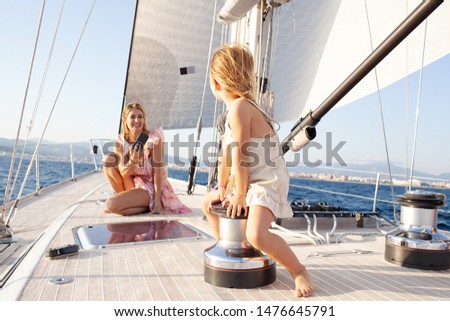 Beautiful rich mother and child daughter enjoying luxury sailing summer holiday trip, using smartphone taking pictures at sunset, smiling outdoors. Family on yacht vacation, aspirational lifestyle.