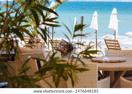 Natural wooden chairs tables empty sunglasses nobody sun beds closed umbrellas flowers on first line beach family restaurant cafe bar aquamarine blue sea ocean sky view morning without visitors guests Royalty-Free Stock Photo #1476648185
