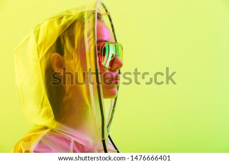 Fashion close up portrait of an attractive young asian woman with long wet hair standing isolated over yellow background, posing in transparent raincoat and sunglasses