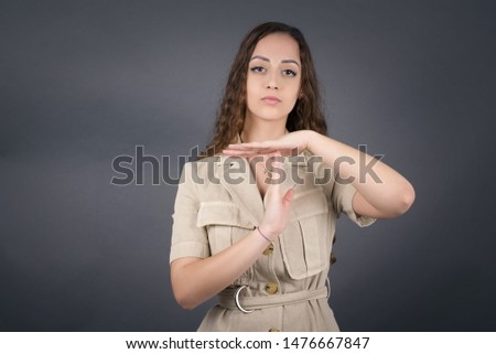 Young pretty blonde girl tired and bored, making a timeout gesture, needs to stop because of work stress, time concept. Standing against gray wall.