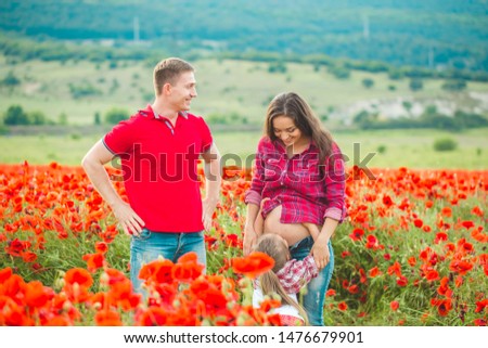 A future mom in a checkered red shirt and jeans walks with her daughter and husband in a poppy field among flowers.