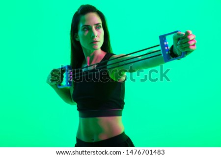 Beautiful confident young fitness girl sitting on a fitness mat isolated over green neon background, working out with an expander