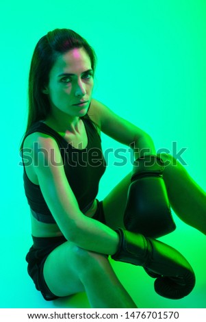 Beautiful confident young fitness girl wearing boxing gloves sitting isolated over green neon background, looking up