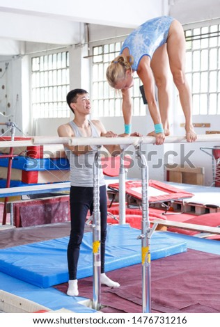 Sporty female gymnast in bodysuit during workout at bars and man helping