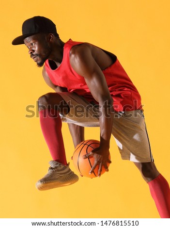 Sport portrait of black man in red shirt and grey shorts playing basketball. Studio, yellow background