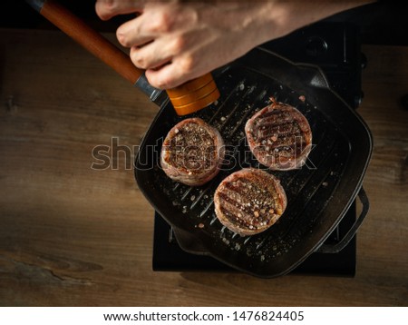 Close up chef hands adding pepper in mill during cooking beef steak on grill pan black background flat lay view.