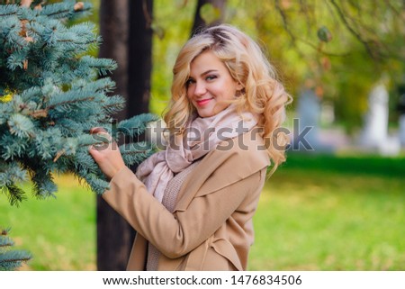 Beautiful elegant blonde woman dressed in a coat standing under the fir-tree in autumn park. Sunny october day.