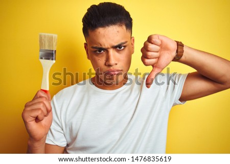 Young brazilian painter man holding brush standing over isolated yellow background with angry face, negative sign showing dislike with thumbs down, rejection concept