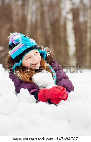 Laughing rosy girl in jacket, knitted hat and mittens lies on snowdrift