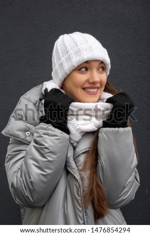 Young, pretty woman in a white knitted hat and scarf dressed in pink jacket against gray wall background. Youth, healthy lifestyle, dreams.